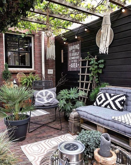 a boho summer patio with a brick floor, layered rugs, a pallet loveseat, a rattan table, potted greenery and candle lanterns and macrame decor