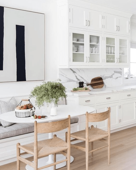 a chic white kitchen with a comfy dining nook, a banquette seating, wooden chairs and an oval table