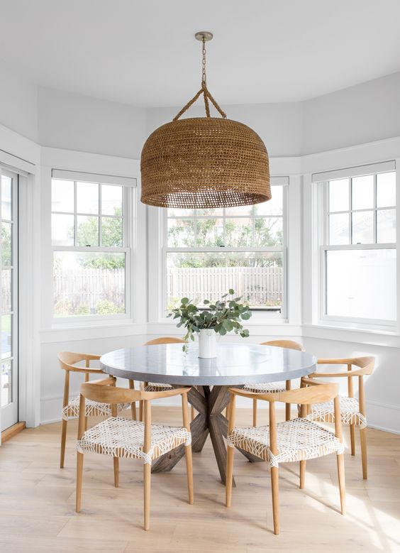 a cool neutral modern boho dining room with a round table, woven chairs, a large woven pendant lamp and greenery in the vase