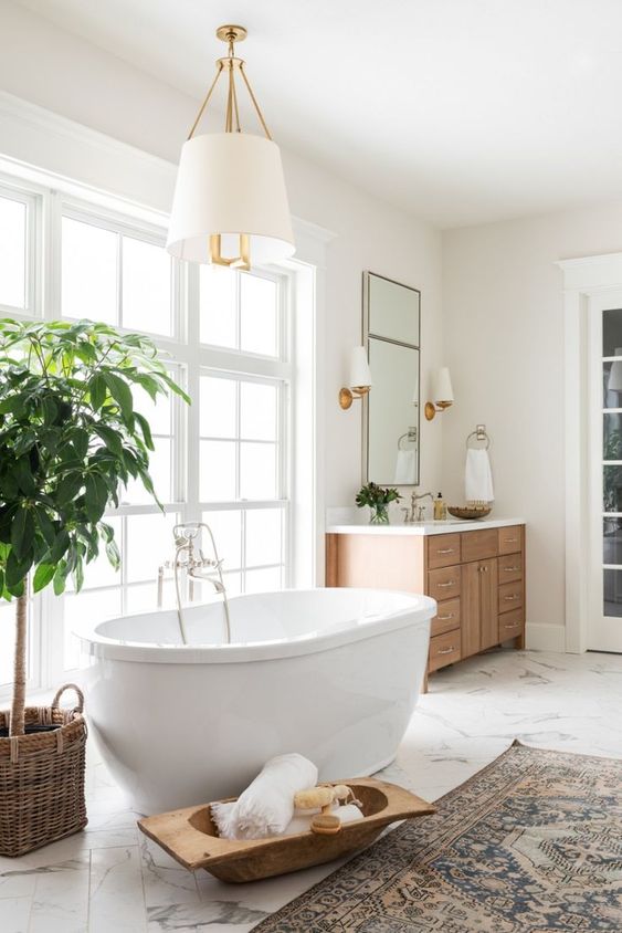 a cozy neutral bathroom with a marble floor, an oval tub, a wooden vanity and a potted tree plus elegant lamps