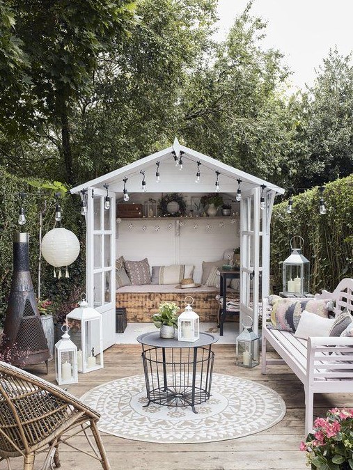 a cozy small terrace with a hearth, a white bench with lots of pillows, a coffee table, a rattan chait, lots of lanterns and a small shed with a daybed