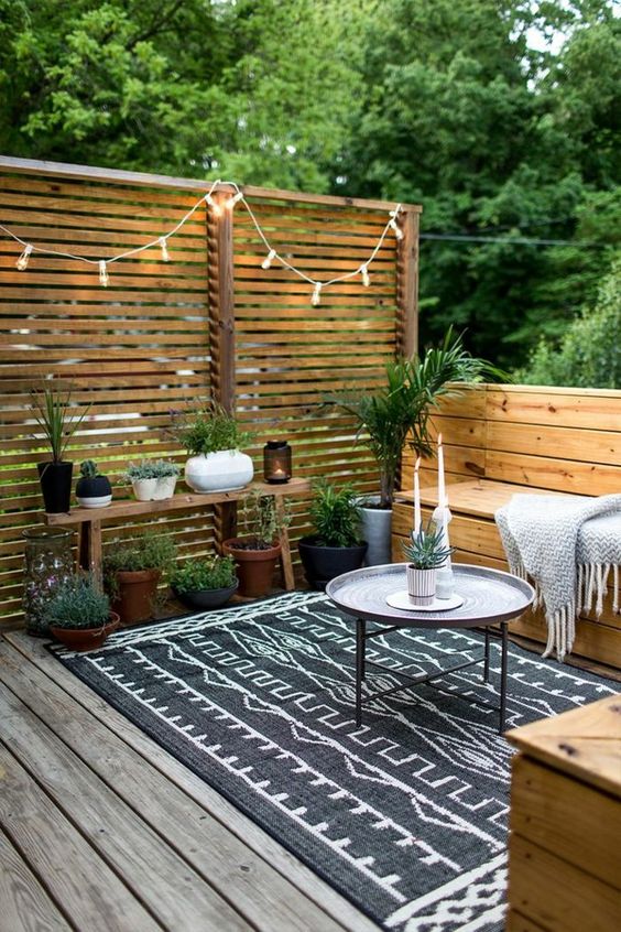 a cozy small terrace with wooden furniture, a small coffee table, a boho rug, potted greenery and lights