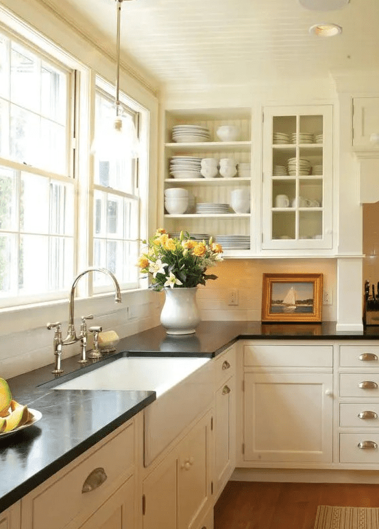 a creamy vintage kitchen with shaker and glass cabinets, black quartz countertops, a window as a backsplash and knobs and handles