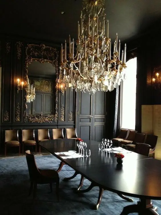 A jaw dropping Gothic dining room with black and gold paneled walls, a large mirror, an oval table, upholstered chairs and a fantastic chandelier