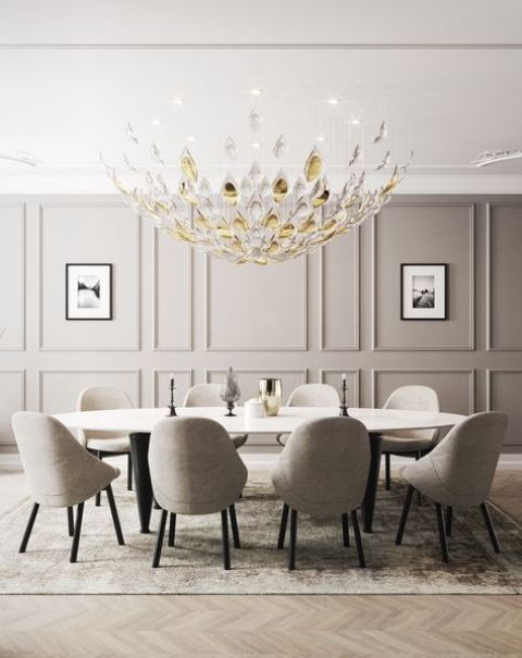 a luxurious dining space with a grye paneled wall, a large beautiful chandelier, a long oval table and grey upholstered chairs