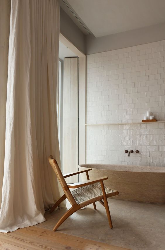 a minimalist yet warm bathroom with glossy white tiles, a wooden bathtub, a rattan chair and neutral curtains on the windows