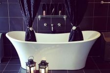 a modern Gothic bathroom with black tiles, a white tub with printed black curtains, a glass shelf and coffin-shaped niches