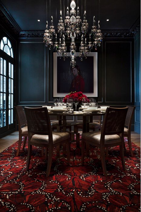 a modern Gothic dining room with black paneled walls, a glass table and upholstered chairs, a red carpet and a crystal chandelier