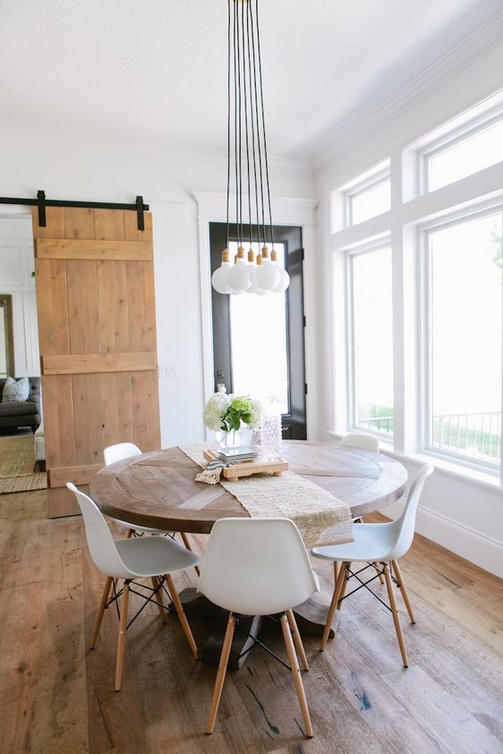 a modern rustic dining room with a round table, white chairs, a cluster of pendant lamps and greenery plus lots of natural light