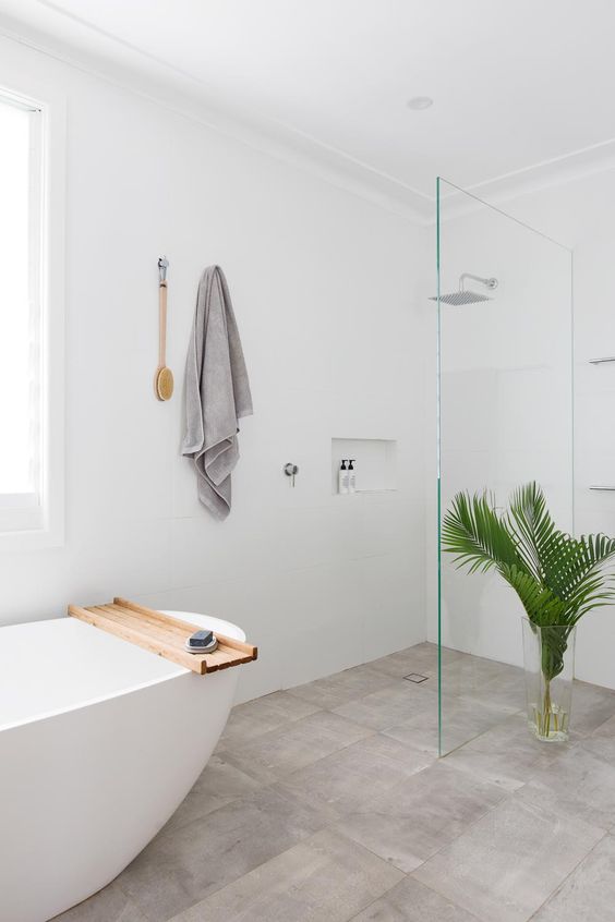 a neutral bathroom clad with tan tiles, an oval tub, neutral textiles and leaves in a vase and an airy glass divider