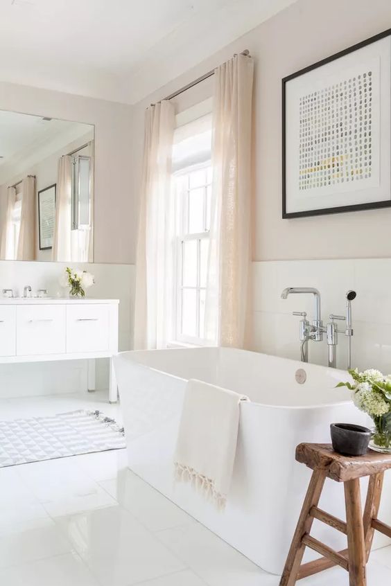 a neutral bathroom with a white vanity and an oval tub, a wooden stool, blush curtains and a bold graphic artwork