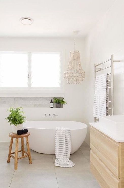 a neutral bathroom with an oval tub, a floating wooden vanity, a beaded chandelier, potted plants and a wooden stool