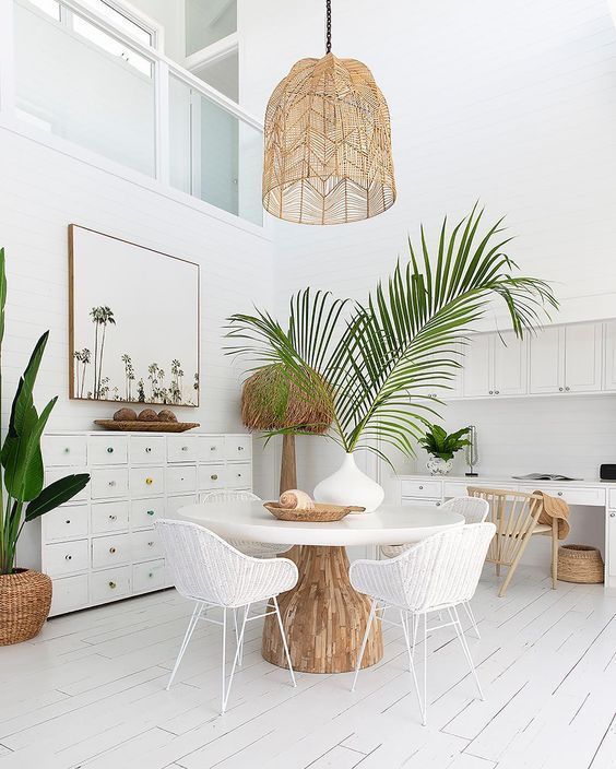 a neutral beachy dining room with a dress, a round table, a woven lamp, white woven chairs, potted plants  and some beachy artworks