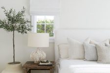 a neutral bedroom with a white upholstered bed, neutral pillows, a white lamp and white shades plus a statement plant