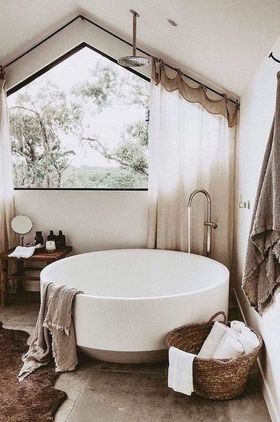 A neutral sabi sabi bathroom with tiles on the floor, a round tub, a basket with towels, a wooden stool and curtains on the window