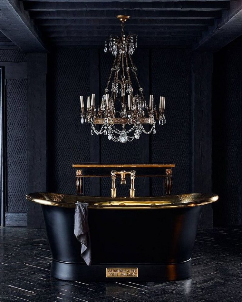 a refined Gothic bathroom with textural graphite grey walls, a black and gold polished bathtub, a crystal chandelier and a black parquet floor