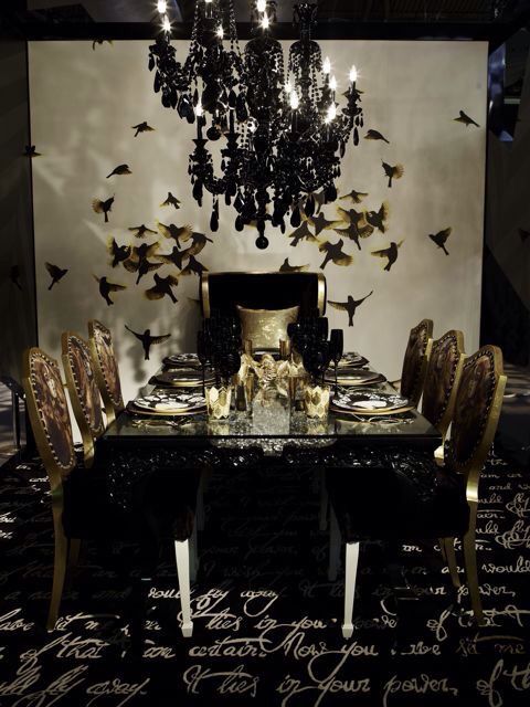 a refined Gothic dining room with a white accent wall with birds, a glass table, gold chairs, several black crystal chandeliers