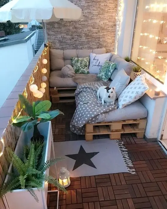 a small and cozy balcony with a pallet L-shaped sofa, printed pillows, plants in pots, lights and candle lanterns