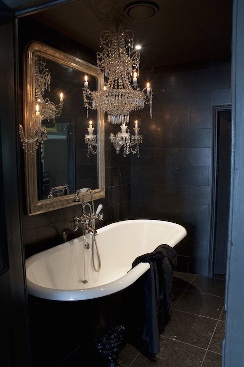 a small and exquisite Gothic bathroom with matte black tiles and a grey tile floor, a vintage black and white tub, a mirror in a gilded frame and a crystal chandelier