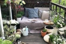 a small and fresh terrace with potted greenery and blooms, a bench with blankets and pillows plus a watering can