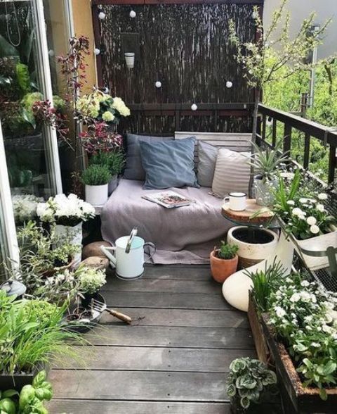a small and fresh terrace with potted greenery and blooms, a bench with blankets and pillows plus a watering can