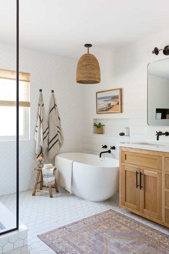 a stylish farmhouse bathroom with mismatching white tiles, a wooden vanity, a woven lamp, an oval tub and black fixtures for a modern feel