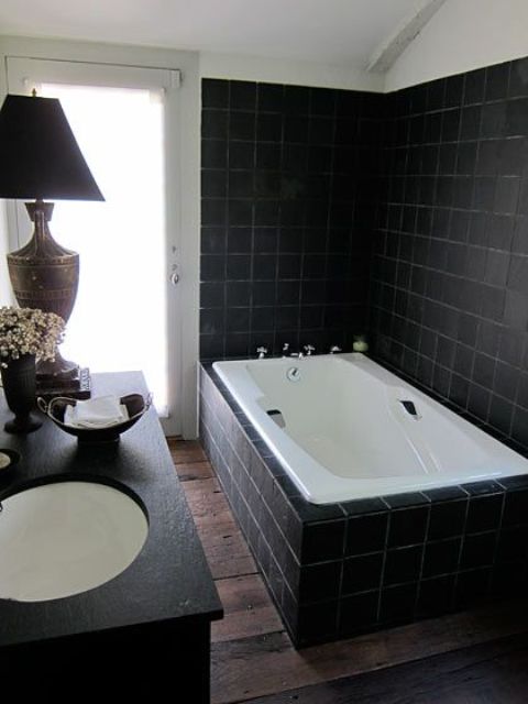 a very laconic Gothic bathroom clad with matte black tiles and a tub clad with them, a black vanity, a chic table lamp and white appliances