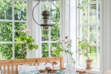 a white Scandi sunroom that is a dining room with stained and painted furniture, with lots of potted blooms and a vintage metal lamp