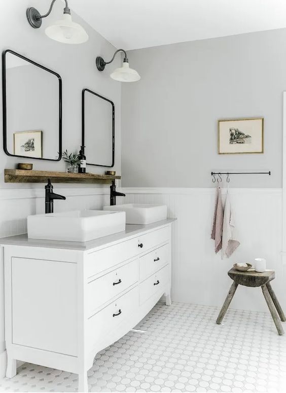 a white and grey farmhouse bathroom with panels, a white vanity and sinks, curved mirrors in black frames and sconces