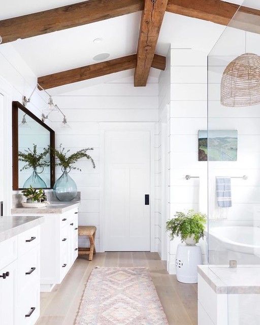 a white coastal bathroom with stained beams on the ceiling, white vanities, a bathtub and a shower space plus a large mirror