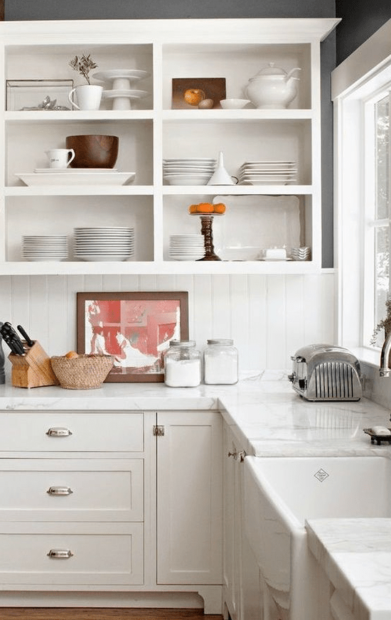 a white cottage kitchen with shaker cabinets and open upper ones, a white beadboard backsplash and white stone countertops