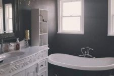 an elegant Gothic bathroom with a black and white tiled floor, a vintage white vanity, a black vintage bathtub, a crystal chandelier and a mirror in a lovely frame