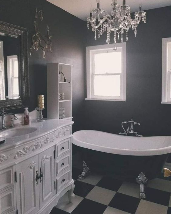 an elegant Gothic bathroom with a black and white tiled floor, a vintage white vanity, a black vintage bathtub, a crystal chandelier and a mirror in a lovely frame