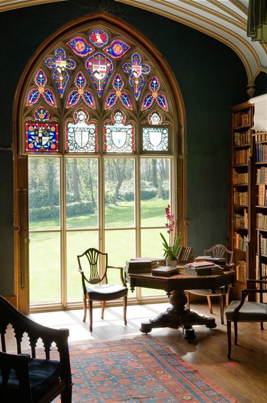 a Gothic-styled home office with an arched mosaic window, dark stained heavy furniture, a large bookcase is a stylish space