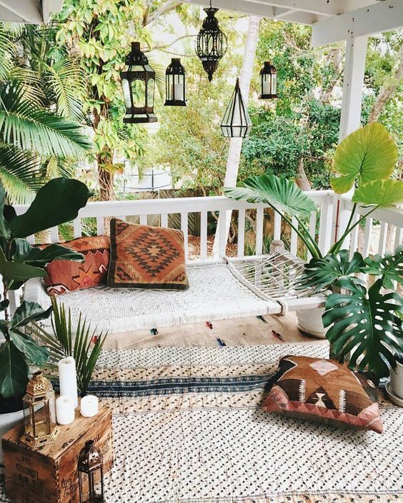 a cool boho porch with a woven hammock, printed textiles, potted greenery, candle lanterns and a wooden chest