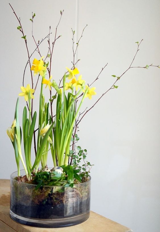 a glass bowl with fake eggs and greenery, daffodils and blooming branches is a modern and chic spring centerpiece