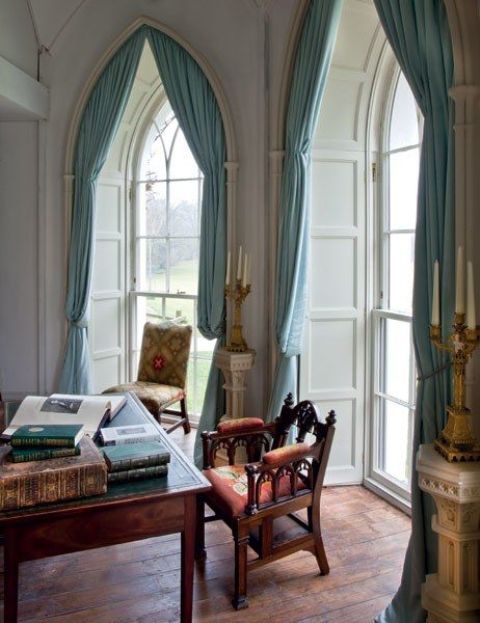 A light colored home office with arched windows, blue curtains, a dark stained desk and heavy chairs plus candles