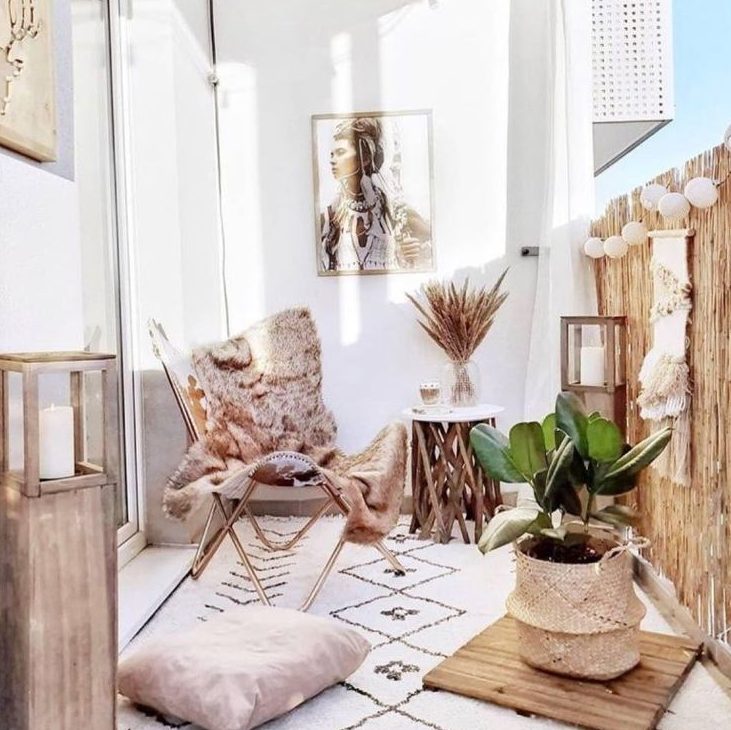 A light filled boho balcony with a folding chair, boho rugs and pillows, a side table with branch legs, baskets and candle lanterns, paper lamps and macrame