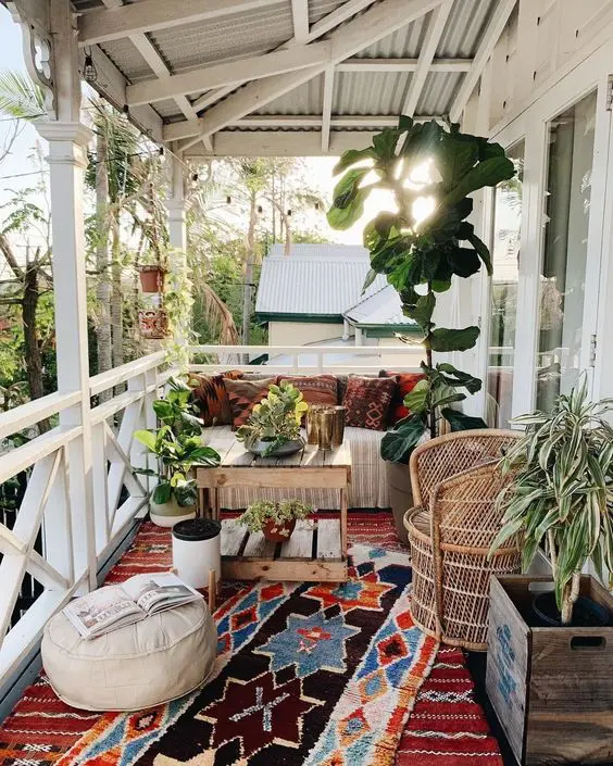 a lovely boho porch with wooden and woven furniture, colorful printed textiles, potted plants and greenery, layered rugs