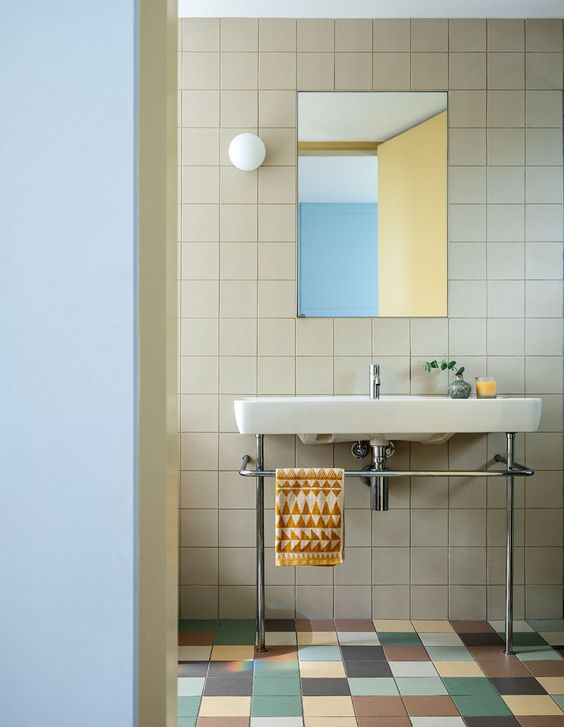 A modern bathroom clad with tan tiles and with matching but colorful tiles on the floor, with a free standing sink and a rectangular mirror