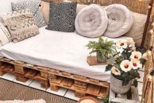 a neutral boho balcony with a pallet daybed, neutral pillows with tassels, potted plants and blooms, candles in candleholders and layered rugs