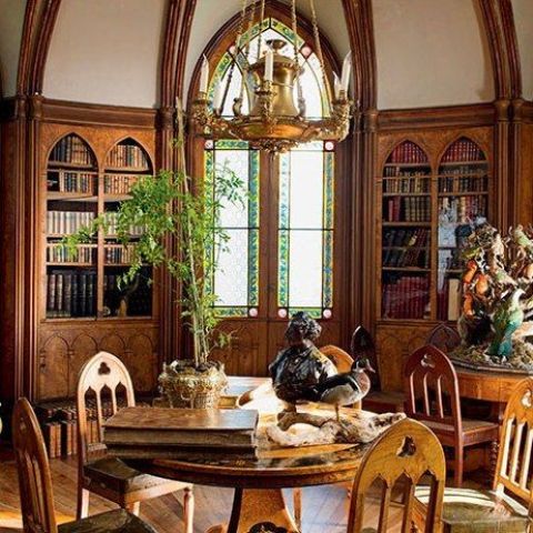a neutral-colored Gothic home library with arched mosaic windows, arched built-in bookcases, round tables and refined chairs plus a gilded chandelier