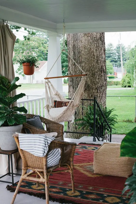 a relaxed boho porch with woven furniture, a hanging macrame chair, boho rugs and potted greenery