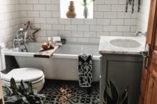 a small boho bathroom with a mosaic tile floor, grey and white tile walls, a tub, potted greenery and baskets for storage
