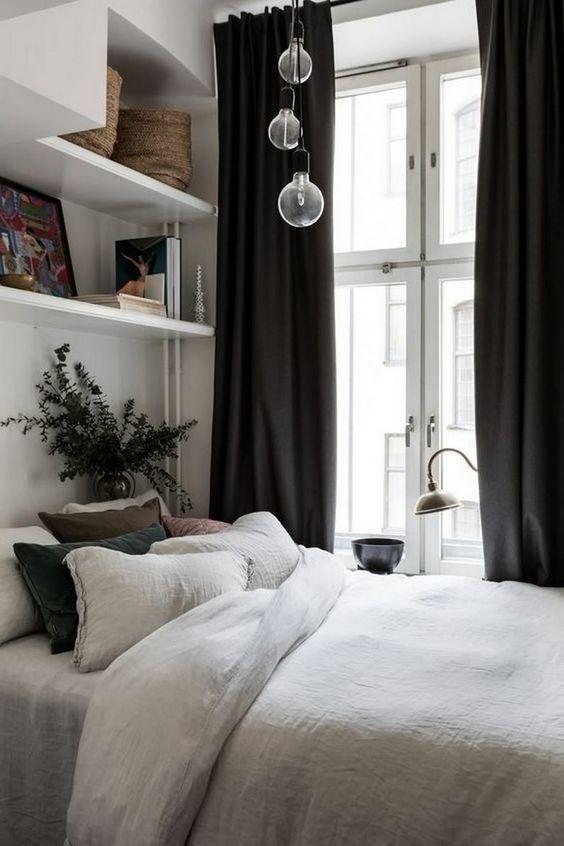 a small chic bedroom with open and closed shelves, a bed with cool bedding, pedant lamps and black curtains