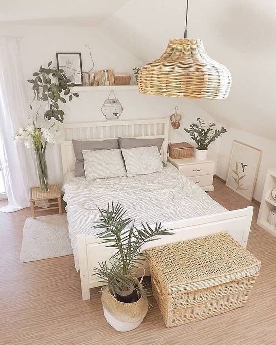 a small neutral bedroom with white furniture, a wicker lamp and a chest, potted plants and blooms and a ledge with artworks