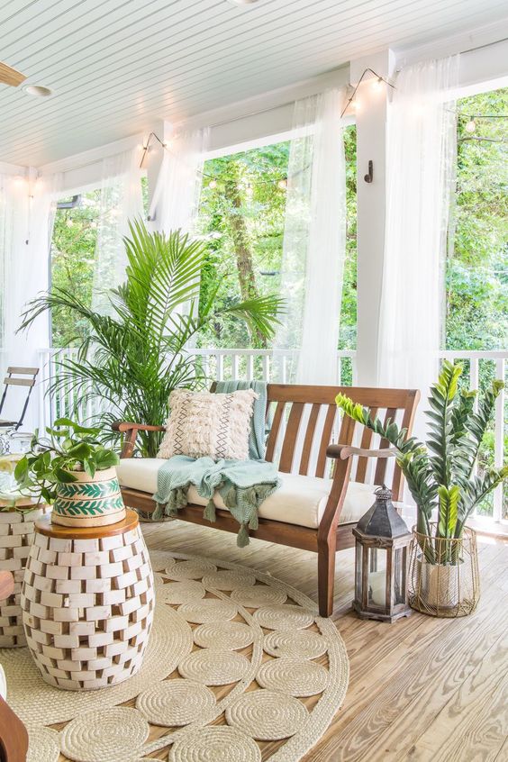 a tropical boho porch with wooden furniture, jute rugs, lots of potted greenery and candle lanterns