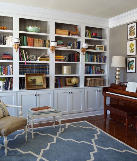 A vintage room with lots of built in shelves, a piano, a vintage chair and ottoman and wall lamps