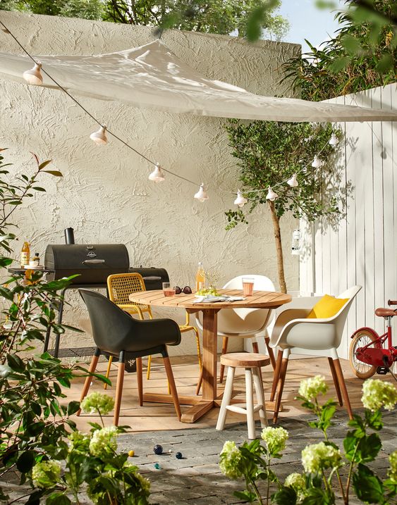 a welcoming sprign terrace with a modern wooden table and mismatching chairs, potted greenery and blooms and some lights over the space