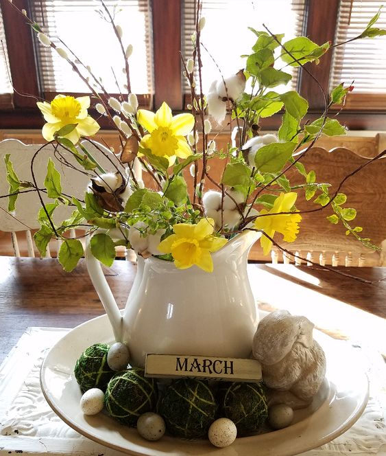 an amazing spring centerpiece of a bowl with moss balls and fake eggs, a jug with greenery, willow and daffodils and cotton
